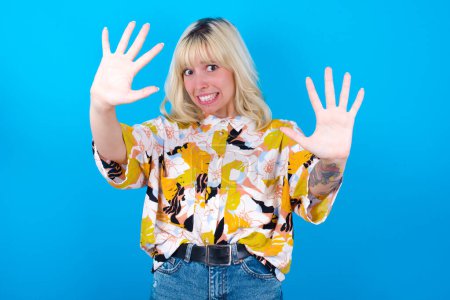 Photo for Dissatisfied Caucasian girl wearing floral shirt isolated over blue background frowns face, has disgusting expression, shows tongue, expresses non compliance, irritated with somebody. - Royalty Free Image