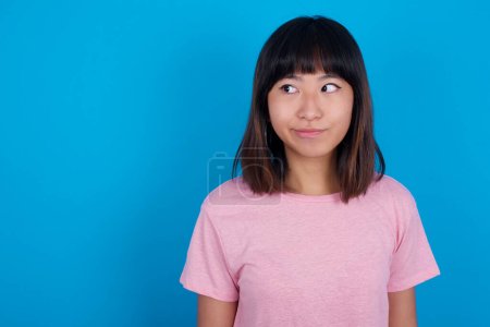Téléchargez les photos : Incroyable perplexe jeune femme asiatique portant t-shirt rose sur fond bleu courbes lèvres et a regard inquiet, voit quelque chose d'horrible devant. - en image libre de droit