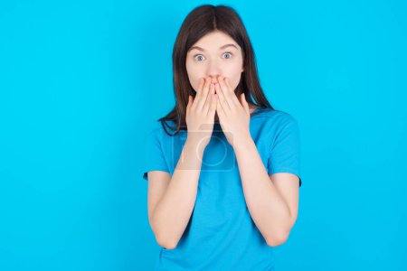 Photo for Young caucasian girl wearing blue t-shirt isolated over blue studio background keeps hands on mouth, looks with eyes full of disbelief, being puzzled with amount of work - Royalty Free Image