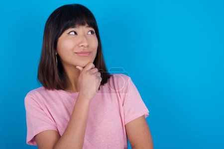 Foto de Joven mujer asiática vistiendo camiseta rosa sobre fondo azul con la mano bajo la barbilla y mirando hacia los lados con expresión dudosa y escéptica, sospechoso y duda. - Imagen libre de derechos