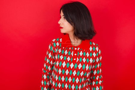 Foto de De cerca foto de perfil lateral morena mujer caucásica con suéter de Navidad sobre fondo rojo no sonriendo atento escuchar concentrado - Imagen libre de derechos