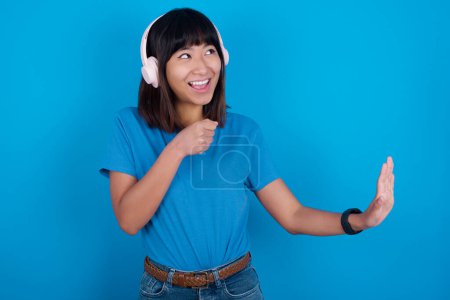 Téléchargez les photos : Heureuse jeune femme asiatique portant un t-shirt bleu sur fond bleu chante chanson préférée garde la main près de la bouche comme si le microphone portait un casque sans fil, écoute de la musique - en image libre de droit