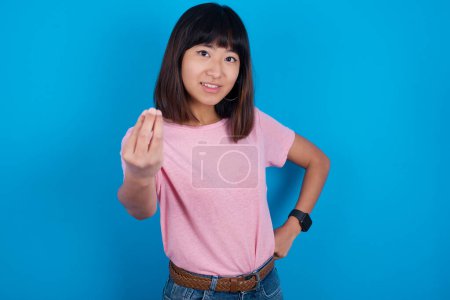 Foto de What the hell are you talking about. Shot of frustrated young asian woman wearing pink t-shirt against blue background gesturing with raised hand doing Italian gesture, frowning, being displeased and confused with dumb question. - Imagen libre de derechos