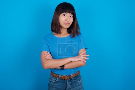 Foto de Confiado joven mujer asiática usando camiseta azul sobre fondo azul con los brazos cruzados mirando a la cámara - Imagen libre de derechos