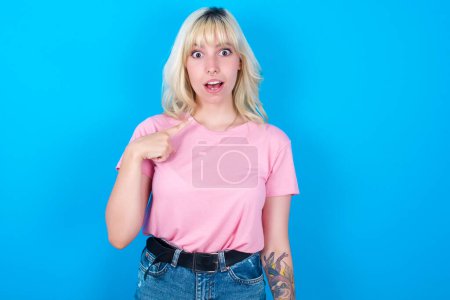 Téléchargez les photos : Caucasienne fille portant rose t-shirt isolé sur fond bleu étant dans la stupeur choqué, a étonné expression pointant vers soi-même avec doigt disant : Qui me? - en image libre de droit