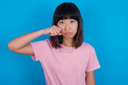 Photo for Unhappy young asian woman wearing pink t-shirt against blue background crying while posing at camera wiping tears with hand. - Royalty Free Image