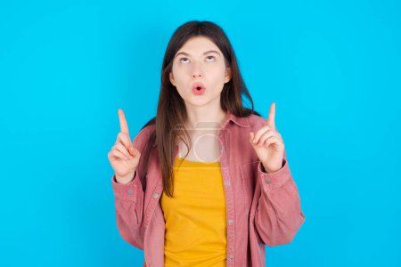 Téléchargez les photos : Jeune fille caucasienne portant chemise rose isolé sur fond bleu étant étonné et surpris regardant et pointant vers le haut avec des doigts montrant quelque chose d'étrange. - en image libre de droit