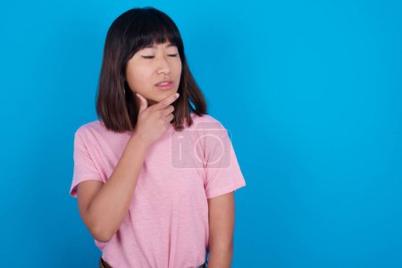 Téléchargez les photos : Prise de vue contemplative réfléchie jeune femme asiatique portant un t-shirt rose sur fond bleu garde la main sous le menton, regarde vers le haut. - en image libre de droit
