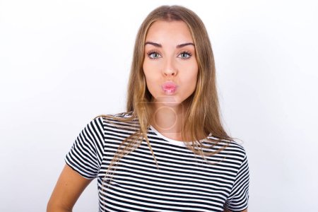 Foto de Retrato de hermosa chica rubia divertida encantadora con camiseta a rayas sobre fondo blanco enviando beso de aire - Imagen libre de derechos