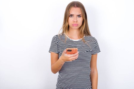 Foto de Molesto chica rubia hermosa insatisfecha con camiseta a rayas sobre fondo blanco utiliza la aplicación de software móvil y la información surrfs en Internet, sostiene la mano móvil moderna - Imagen libre de derechos
