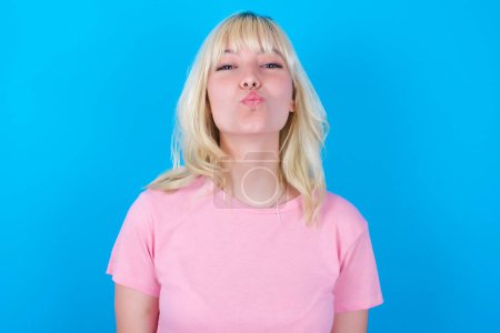 Photo for Caucasian girl wearing pink t-shirt isolated over blue background keeps lips as going to kiss someone, has glad expression, grimace face. Standing indoors. Beauty concept. - Royalty Free Image