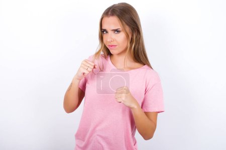 Téléchargez les photos : Portrait d'attrayant jeune fille caucasienne portant T-shirt rose sur fond blanc tenant la main devant lui en position de boxe va se battre. - en image libre de droit