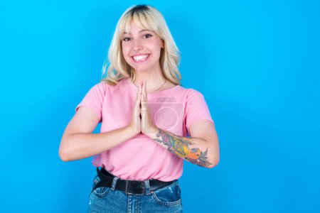 Photo for Caucasian girl wearing pink t-shirt isolated over blue background keeps palms together, has pleased expression. Glad attractive male makes request, pleads for mercy. Hopeful young adult. - Royalty Free Image