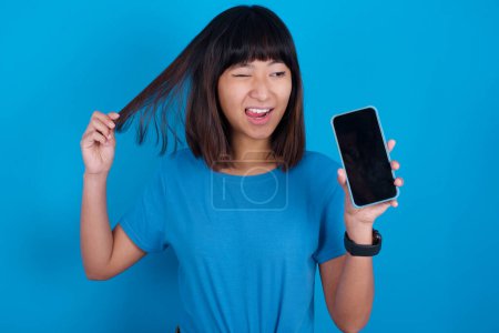 Téléchargez les photos : Photo de belle jolie jeune femme asiatique portant t-shirt bleu sur fond bleu démontrer téléphone écran tenir des queues de cheveux - en image libre de droit