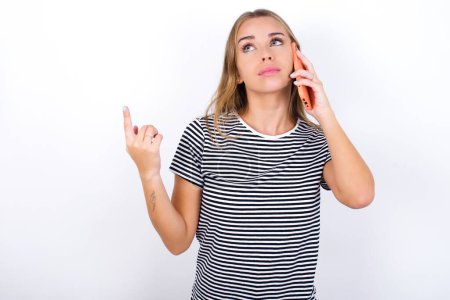 beautiful blonde girl wearing striped t-shirt on white background speaks on mobile phone spends free time indoors calls to friend.