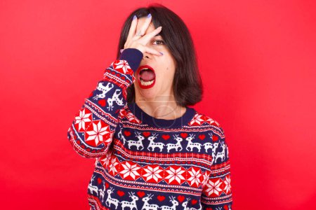 Foto de Morena mujer caucásica vistiendo suéter de navidad sobre fondo rojo asomándose en shock cubriendo la cara y los ojos con la mano, mirando a través de los dedos con expresión avergonzada. - Imagen libre de derechos