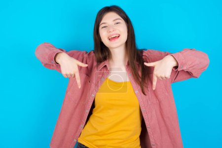 Téléchargez les photos : Jeune fille caucasienne portant chemise rose isolé sur fond bleu avec une expression positive, pointe vers le bas avec les deux index, a large sourire intéressé. Regardez, s'il vous plaît.. - en image libre de droit