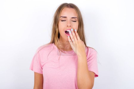 Photo for Sleepy Young Caucasian girl wearing pink T-shirt on white background yawning with messy hair, feeling tired after sleepless night, yawning, covering mouth with palm. - Royalty Free Image
