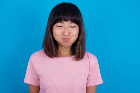 Téléchargez les photos : Jeune femme asiatique portant un t-shirt rose sur fond bleu joues gonflées avec un visage drôle. Bouche gonflée avec de l'air, expression folle. - en image libre de droit