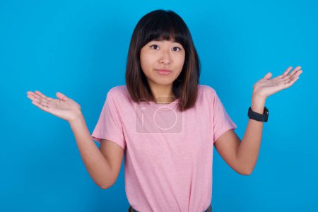 Téléchargez les photos : Jeune femme asiatique perplexe et désemparée portant un t-shirt rose sur fond bleu avec les bras ouverts, haussant les épaules, disant : qui s'en soucie, alors quoi, je ne sais pas. - en image libre de droit