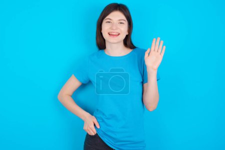 Foto de Joven chica caucásica vistiendo camiseta azul aislada sobre fondo estudio azul renunciando a decir hola o adiós feliz y sonriente, gesto de bienvenida amistoso. - Imagen libre de derechos
