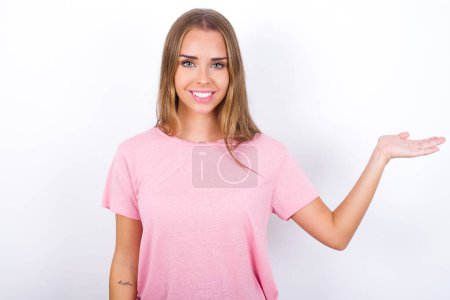 Téléchargez les photos : Positif heureux Jeune fille caucasienne portant T-shirt rose sur fond blanc dit : wow combien il est passionnant, a une expression étonnante, montre quelque chose sur l'espace vide avec la main ouverte. Concept de publicité. - en image libre de droit