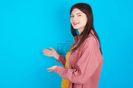 Foto de Joven chica caucásica con camisa rosa aislado sobre fondo azul Invitar a entrar sonriendo natural con las manos abiertas. Señal de bienvenida. - Imagen libre de derechos