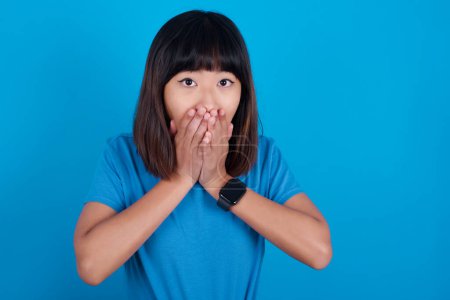 Foto de Joven asiático mujer usando azul camiseta contra azul fondo mantiene las manos en la boca, mira con los ojos llenos de incredulidad, siendo desconcertado con la cantidad de trabajo - Imagen libre de derechos