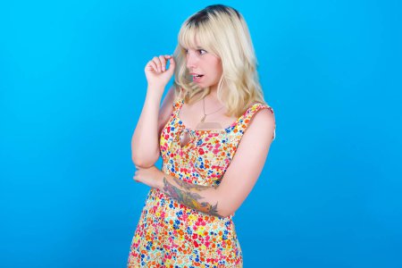 Photo for Astonished caucasian girl wearing floral dress isolated over blue background looks aside surprisingly with opened mouth. - Royalty Free Image