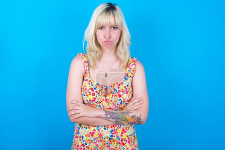 Photo for Gloomy dissatisfied caucasian girl wearing floral dress isolated over blue background looks with miserable expression at camera from under forehead, makes unhappy grimace - Royalty Free Image