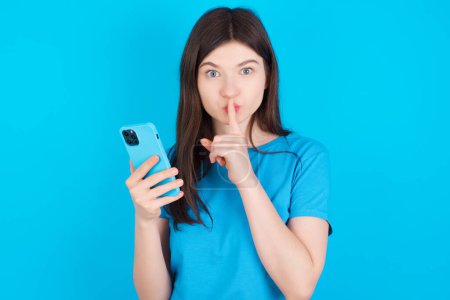 Foto de Joven caucásico chica usando azul camiseta aislado sobre azul estudio fondo celebración moderno gadget pedir no contar secretos - Imagen libre de derechos