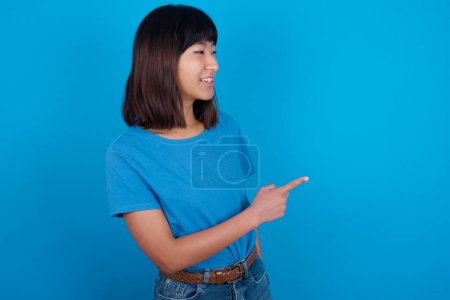 Foto de Feliz alegre sonriente joven asiática mujer con camiseta azul sobre fondo azul mirando y señalando a un lado con la mano. Copiar espacio y concepto de publicidad. - Imagen libre de derechos