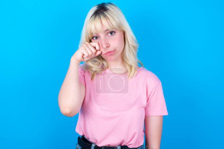 Téléchargez les photos : Malheureux caucasien fille portant rose t-shirt isolé sur fond bleu pleurer tout en posant à caméra fouetter larmes avec main. - en image libre de droit