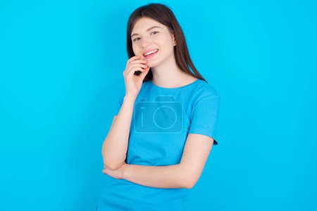 Téléchargez les photos : Insouciant succès jeune fille caucasienne portant bleu t-shirt isolé sur fond studio bleu touchant mâchoire regardant caméra inclinaison tête souriant dents blanches ravi. Concept de soins dentaires. - en image libre de droit