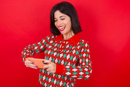 Téléchargez les photos : Brune caucasienne femme portant chandail de Noël sur fond rouge tenant dans les mains cellulaire jouer à des jeux vidéo ou bavarder - en image libre de droit