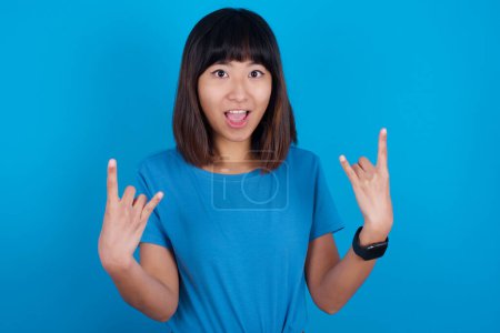 Téléchargez les photos : Jeune femme asiatique portant t-shirt bleu sur fond bleu fait signe rock n roll semble confiant et joyeux aime la musique cool à la fête. Concept de langage corporel. - en image libre de droit