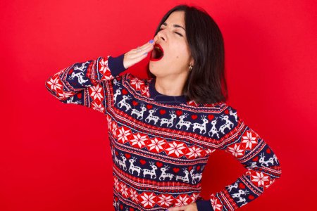 Téléchargez les photos : Brune caucasienne portant un pull de Noël sur fond rouge étant fatigué et bâillant après avoir passé toute la journée au travail. - en image libre de droit