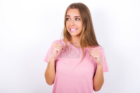 Photo for Young Caucasian girl wearing pink T-shirt on white background clenches fists and awaits for something nice happened looks away bites lips and waits announcement of results - Royalty Free Image