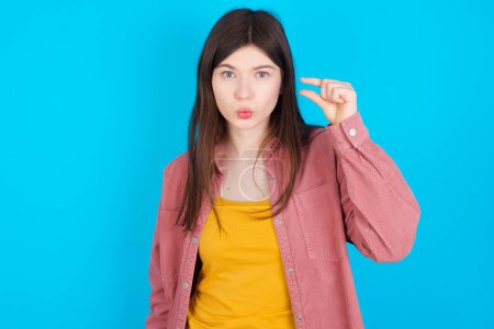 Photo for Shocked young caucasian girl wearing pink shirt isolated over blue background shows something little with hands, demonstrates size, opens mouth from surprise. Measurement concept. - Royalty Free Image