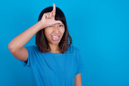 Foto de Divertido joven asiático mujer usando azul camiseta contra azul fondo hace perdedor gesto burlándose de alguien palos fuera lengua haciendo mueca cara. - Imagen libre de derechos