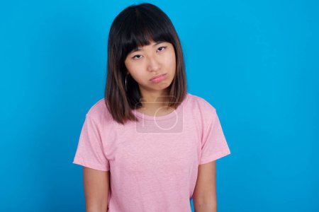 Photo for Gloomy, bored young asian woman wearing pink t-shirt against blue background frowns face looking up, being upset with so much talking hands down, feels tired and wants to leave. - Royalty Free Image