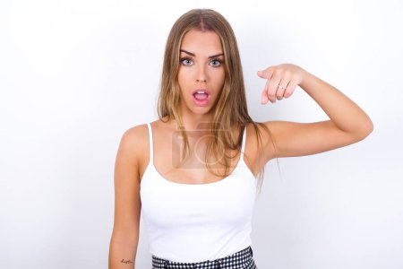 Photo for Shocked Young Caucasian girl wearing white tank top on white background points front with index finger at camera and. Surprise and advertisement concept. - Royalty Free Image
