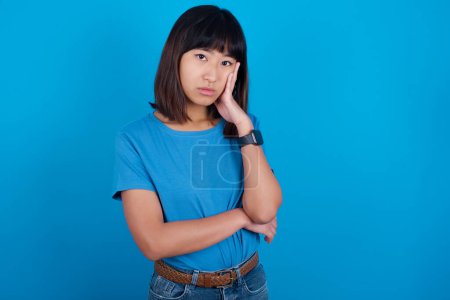 Téléchargez les photos : Très ennuyé jeune asiatique femme portant bleu t-shirt sur fond bleu tenant la main sur la joue tout en le soutenant avec une autre main croisée, l'air fatigué et malade. - en image libre de droit