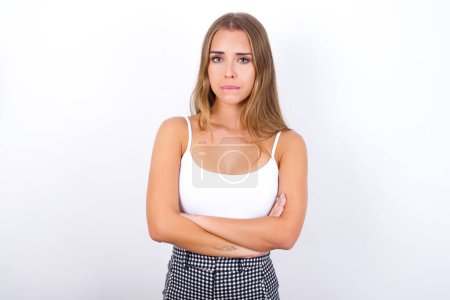 Young Caucasian girl wearing white tank top on white background biting his mouth and looking worried and scared crossing arms, worry and doubt.