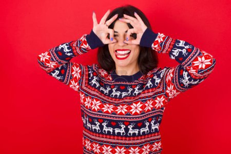 Téléchargez les photos : Brune caucasienne portant un pull de Noël sur fond rouge faisant un geste correct comme des jumelles sortant la langue, les yeux regardant à travers les doigts. Expression folle. - en image libre de droit
