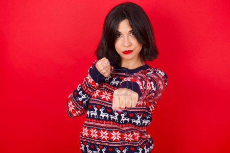 Téléchargez les photos : Femme caucasienne brune portant un pull de Noël sur fond rouge Poing de poing pour se battre, attaque agressive et en colère, menace et violence - en image libre de droit
