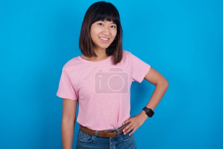 Photo for Studio shot of cheerful young asian woman wearing pink t-shirt against blue background keeps hand on hip, smiles broadly. - Royalty Free Image