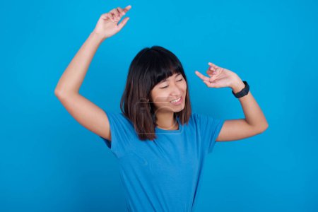 Photo for Photo of upbeat young asian woman wearing blue t-shirt against blue background has fun and dances carefree wear being in perfect mood makes movements. Spends free time on disco party - Royalty Free Image