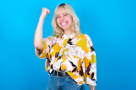 Photo for Overjoyed caucasian girl wearing floral shirt isolated over blue background glad to receive good news, clenching fist and making winning gesture. - Royalty Free Image