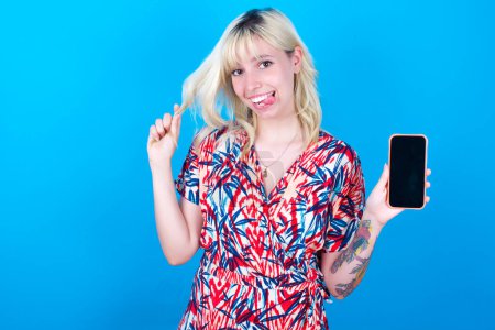 Photo for Photo of nice pretty caucasian girl wearing floral dress isolated over blue background demonstrate phone screen hold hair tails - Royalty Free Image